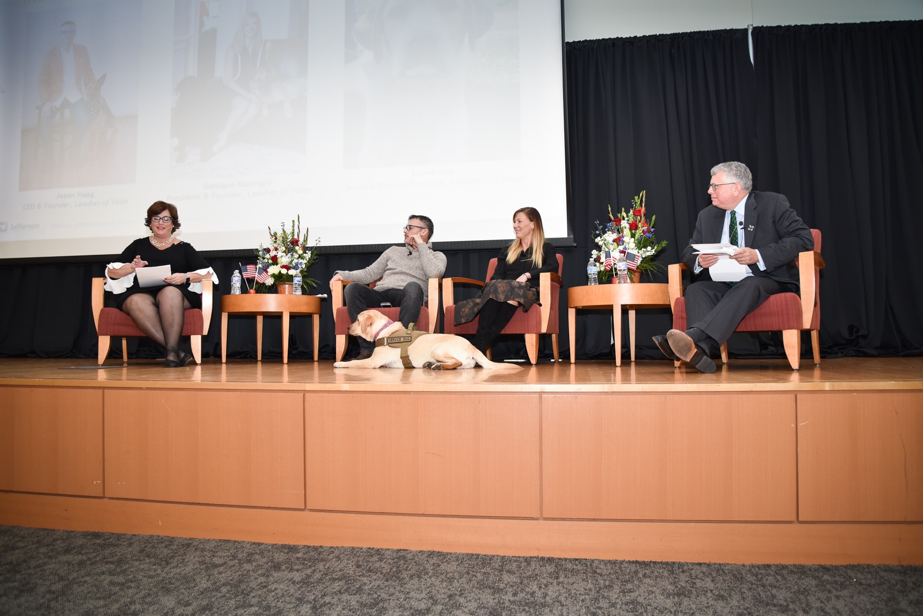 Panelists sit on stage with the dog Maggie on the ground