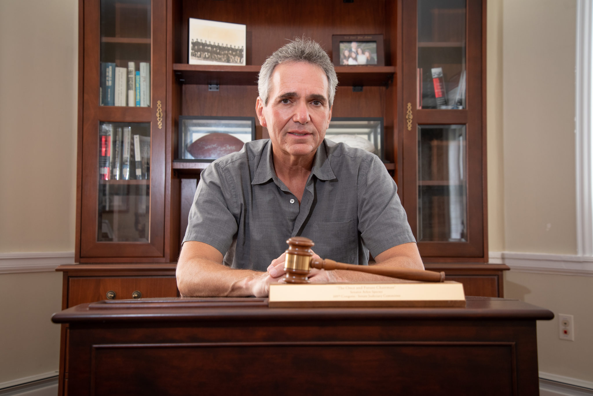 white man in grey shirt sitting at desk with gavel laying on top of a book